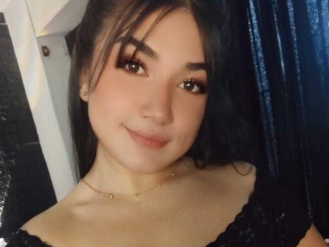 AmyRousse, met zwart haar en bruine ogen, draagt een zwarte top van kant, heeft een schattige glimlach, subtiele make-up en een gouden ketting. Haar borsten zijn gedeeltelijk zichtbaar..