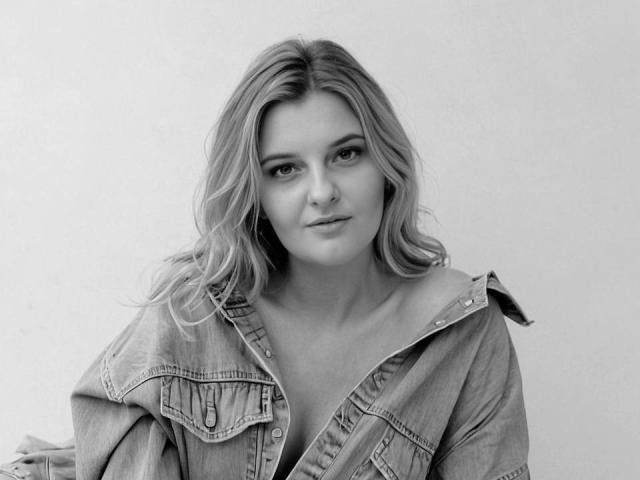 Schwarz-weißes Foto von AssiaS mit welligem, blondem Haar, die eine Jeansjacke trägt und direkt in die Kamera lächelt, wobei ihre Brüste durch die Jacke sichtbar sind..