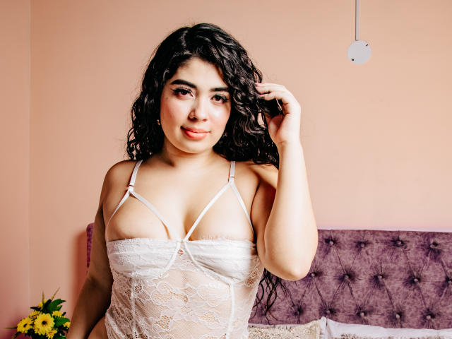 AgathaLynx with curly black hair, wearing a white lace lingerie top, exposing small boobs with thin straps, smiling, right hand in hair, standing in a pink room.