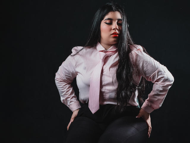 AgathaLynx con lunghi capelli neri, camicia rosa, cravatta rosa, pantaloni neri, mani sui fianchi, che guarda in basso, rossetto rosso, sfondo scuro, formosa, sicura di sé, sexy, glutei visibili, seno delineato..
