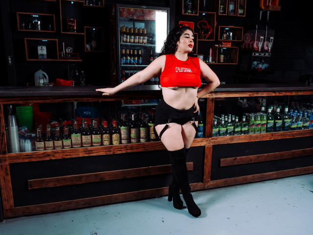 AgathaLynx with curvy body, red crop top, black shorts, garter, and thigh-high boots, leaning on bar counter, red lipstick, dark hair, standing in bar with liquor bottles.