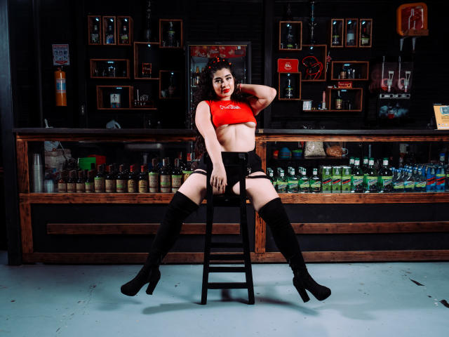AgathaLynx sits on a stool, legs spread, wearing a red crop top, black garter shorts, and thigh-high boots. Her tits are small, with dark nipples, and she's looking at the camera.
