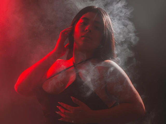 AgathaLynx with red-lit smoke, dark hair, and a black top, holding her chest, showing her tits, with a hand in her hair, looking seductive and confident.
