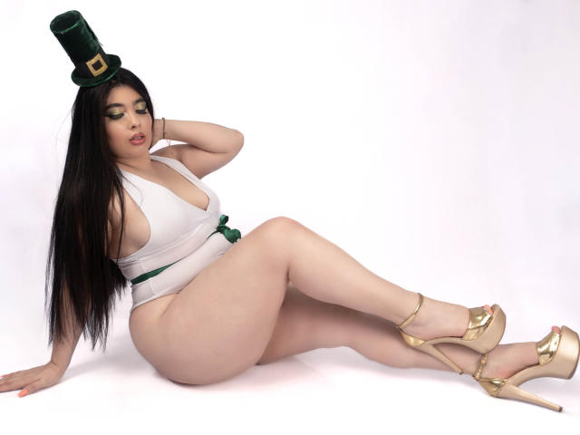 AgathaLynx mit langem, schwarzem Haar, einem weißen Einteiler, einem grünen Zylinderhut, goldenen High Heels, sitzend, mit großem Hintern, einem kleinen grünen Gürtel, einer Hand auf dem Kopf, nach unten schauend, blasser Haut, sexy..