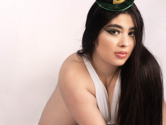 AgathaLynx, con lunghi capelli neri, un cappello di velluto verde, una maglia bianca e un trucco leggero, che mette in mostra il seno, con un'espressione sexy e sicura di sé. Labbra rosa pallido. Sfondo bianco..