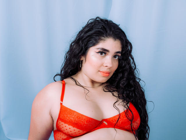 AgathaLynx with long curly black hair, wearing a red lace bra, showing her tits, has a light skin tone, and a subtle smile against a blue curtain.