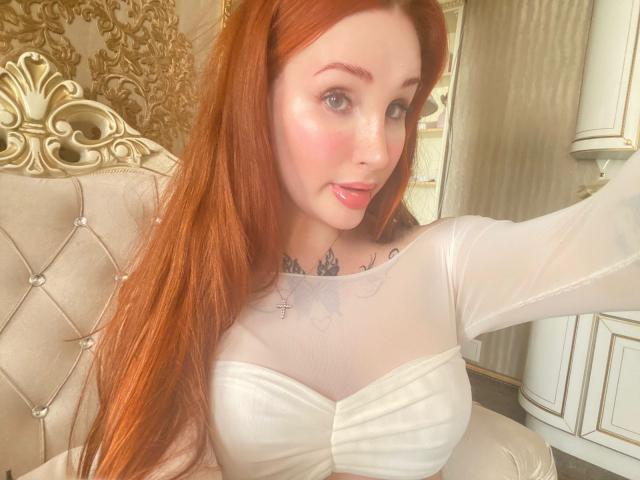 Damen mit langen, roten Haaren, heller Haut und rosafarbenen Lippen, die ein weißes, durchsichtiges Oberteil tragen und ihre großen Brüste und Tattoos zeigen, machen ein Selfie in einem prunkvollen Raum..
