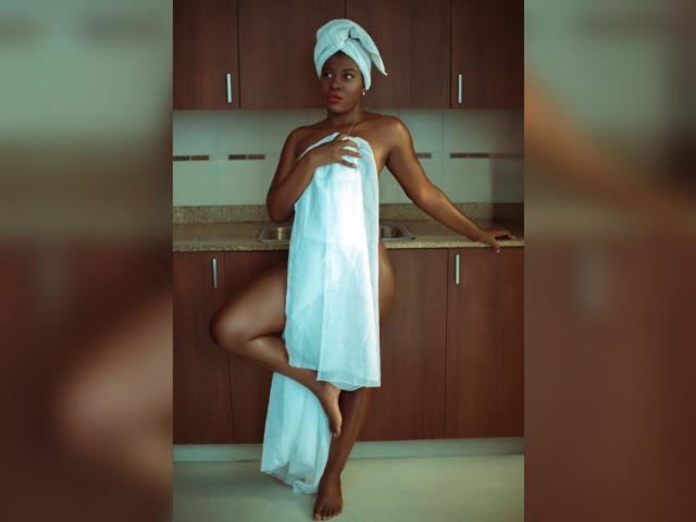 AilynQueen, naked, with white towel covering her tits and pussy, stands in a kitchen, her brown ass and legs visible, a towel on her head, looking at the camera.