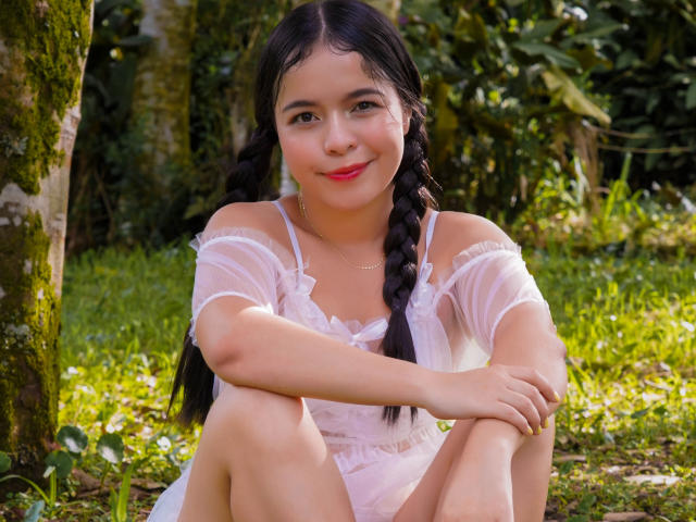 LissyCarter with black braids, sitting on grass, wearing a sheer white dress, smiling, legs up, arms around knees, cute, small tits, pink lipstick, forest background, daytime.