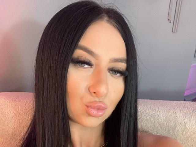 LexyVoss mit langem, schwarzem Haar, vollen, rosafarbenen Lippen und dichten, schwarzen Wimpern, die leicht den Mund verzogen hat. Sie hat einen hellen Hautton und blickt direkt in die Kamera..