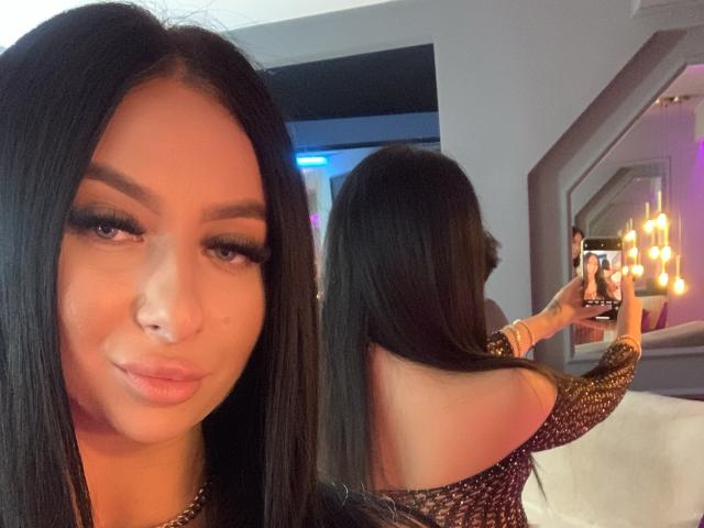 LexyVoss, con cabello negro largo, cejas pobladas y pestañas grandes, se está tomando un selfie en un espejo, vistiendo una blusa brillante con un hombro descubierto, mostrando su espalda y trasero..