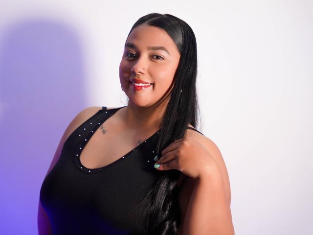 KattySantana with long black hair, smiling, wearing a black tank top with studs, touching her hair, dark skin, small tattoo on chest, red lipstick, green nails.