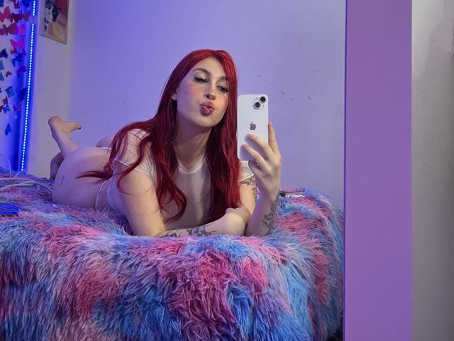 SammyViolet with red hair, pouty lips, lying on blue-pink furry bed, taking selfie with iPhone, wearing white tee, showing tattoo on left arm, bare ass.