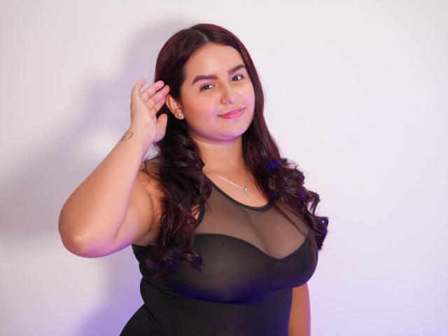 MelodyJames has dark curly hair, a cute smile, and wears a sheer black top showing her big boobs. She's touching her hair with a tattoo on her wrist.