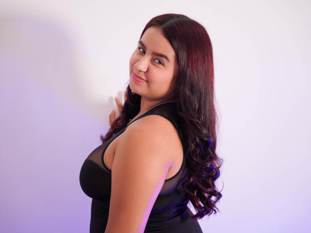 MelodyJames with dark, wavy hair, black sheer top, and curvy, medium-sized tits, smiling at the camera, hand near face, white background, soft purple and blue lighting.