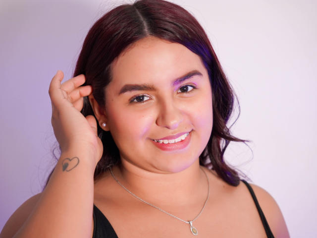 MelodyJames has dark red hair, a small heart tattoo on her right wrist, and is smiling with her hand near her head. She wears a black top and a necklace.