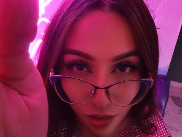 MoollyEvanns, con gli occhiali, il viso illuminato di rosa, i capelli castani e un braccio alzato, che mostra un dito, ha un aspetto carino e seducente, e indossa una maglietta rosa scintillante..