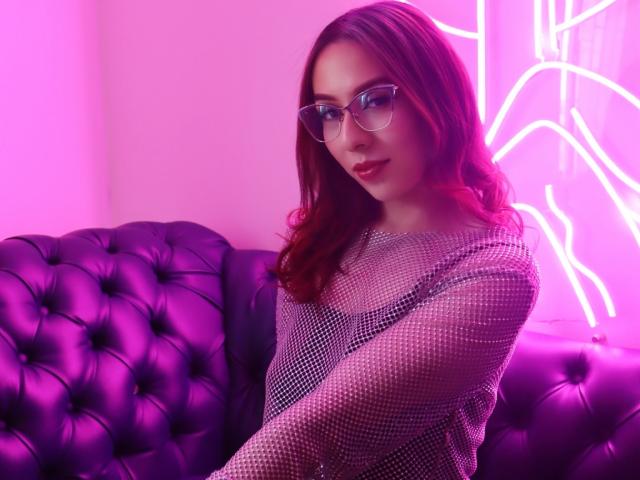 MoollyEvanns, con cabello rojo, gafas y una blusa de malla brillante, sentada en un sofá morado con adornos, con luces de neón rosas de fondo, luciendo sexy y segura de sí misma..