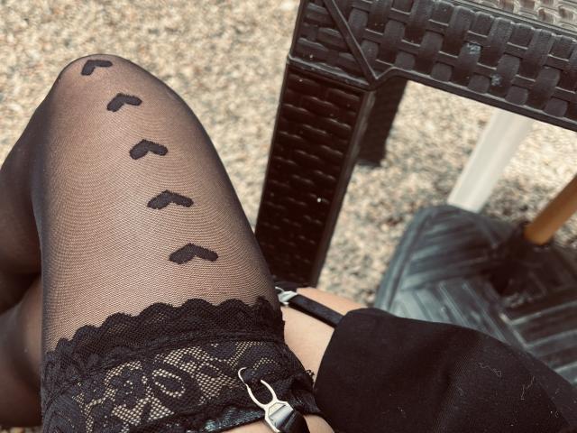 LillyTaylor's black lace-topped stockings with black butterfly patterns, sitting with garter straps visible, black shoes, and a wicker chair in the background.