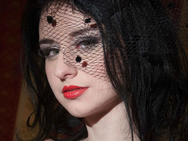 Kathrynne-hot has pale skin, black wavy hair, red lipstick, and black mesh with small holes covering her eyes. She's looking slightly to the side with a subtle smile.