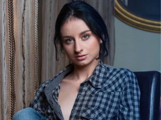 Kathrynne-hot with dark hair, wearing a plaid shirt, slightly open, showing a bit of her tits, sitting on a black chair, looking at the camera with a subtle smile.