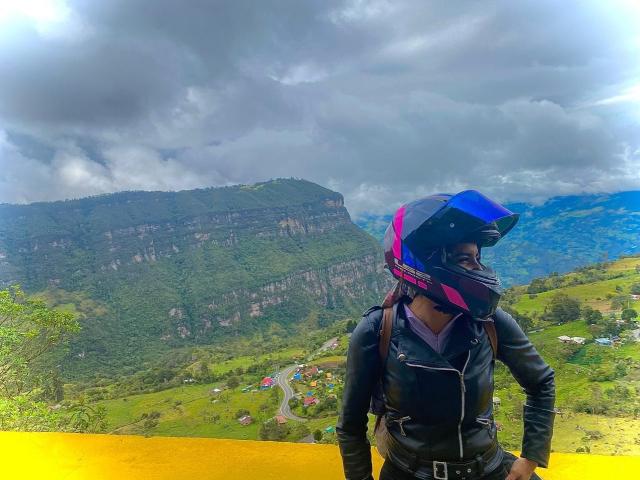 SofyaLane portant une veste en cuir noir, un casque bleu et rose, debout les mains sur une rambarde jaune, avec une montagne verte et un ciel nuageux en arrière-plan. Ses fesses sont légèrement visibles..