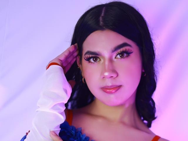 AshleyPiinkk with dark wavy hair, thick black eyebrows, long eyelashes, pink lipstick, and a white shirt with red sleeves, touching her ear. Purple and pink lighting.