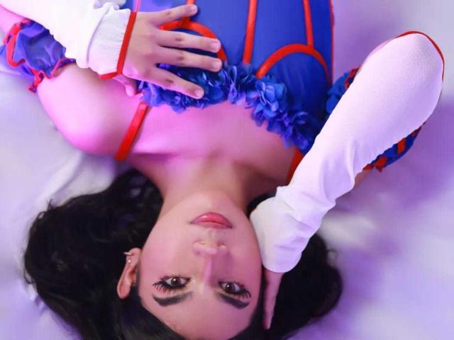AshleyPiinkk lying on her back, wearing a blue and red costume with white sleeves, hands on her chest, dark wavy hair, looking up with a slight smile.