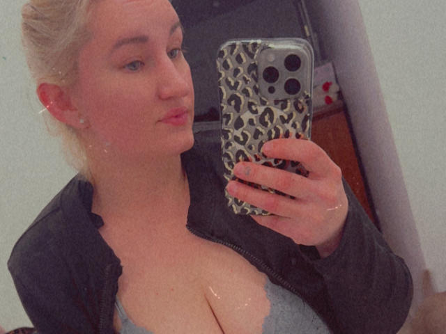 LillieLove taking a selfie with a leopard print phone case, showing her big boobs in a gray lace bra, wearing a black zip-up jacket, with a slight pout.