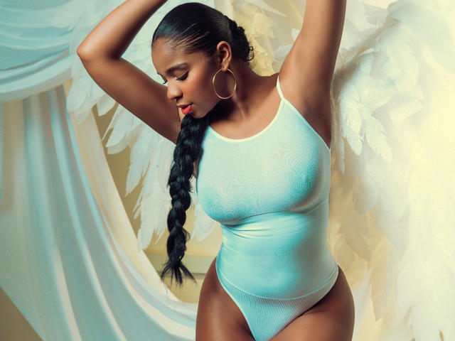 LisaMendez with dark skin, long braided hair, wearing a light blue, textured, sleeveless top, hoop earrings, arms raised, showing her big, round, perky ass and small, firm tits.