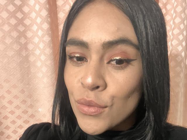 StellaBeckker has dark straight hair, brown eyes with gold eyeliner, and is making a pouty face with slightly pursed pink lips. She has a small nose and is wearing a black top.
