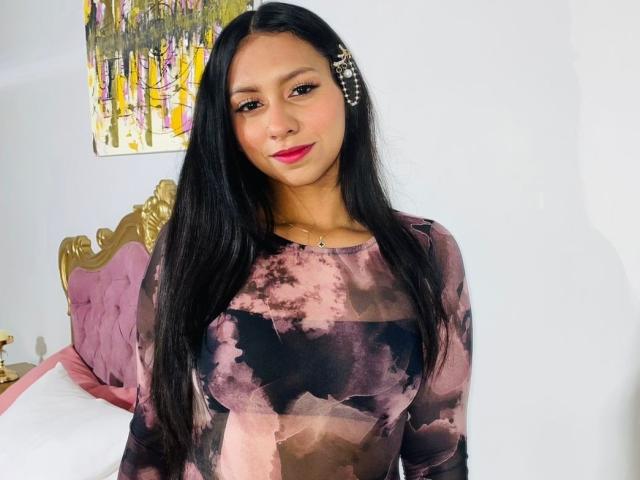 AshleyRosel with long black hair, wearing a pink and black tie-dye top, has small tits and a cute smile, pink lipstick, and a small pearl necklace, standing in a room.