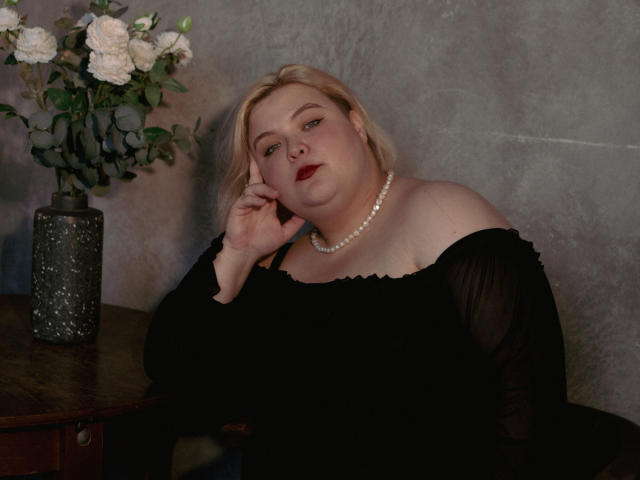 EllaBigBabe, a chubby blonde woman with red lipstick, wears a black off-shoulder dress and pearl necklace, resting her head on her hand, next to a floral vase.