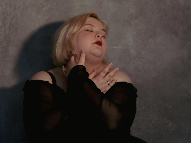EllaBigBabe with blonde bob, eyes closed, red lipstick, black sheer top, hands near face, ring visible, soft shadow, slightly chubby, sexy, sensual expression, light skin, gentle smile.