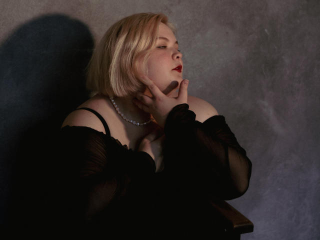 EllaBigBabe with blonde bob, wearing black sheer top, pearl necklace, red lipstick, hand on cheek, looking up, shadow on wall, soft lighting, subtle elegance.