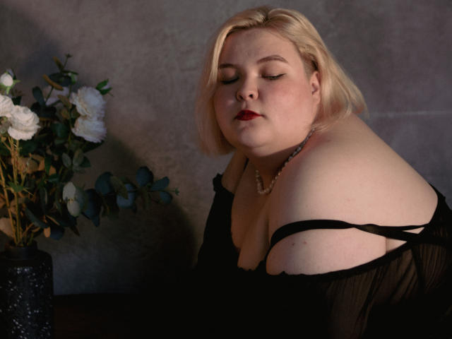 EllaBigBabe with pale skin, blonde bob, red lipstick, black sheer top, off-shoulder, pearl necklace, eyes closed, leaning forward, black vase with white flowers on left.