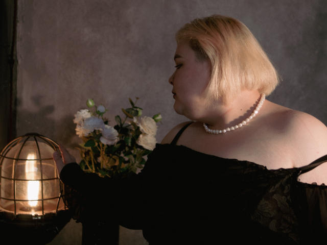 EllaBigBabe with blonde bob, off-shoulder black dress, pearl necklace, holding bouquet of white and green flowers, next to warm globe light. Soft, moody lighting.