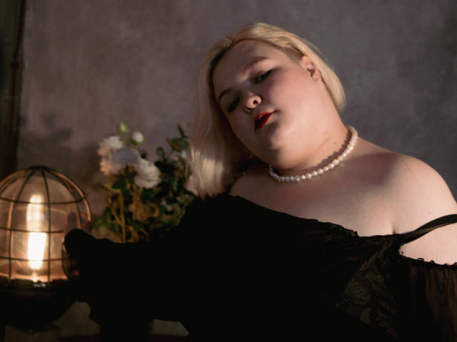 EllaBigBabe with pale skin, blonde hair, and red lipstick, wearing a black lace off-shoulder top and pearl necklace, looking down with a seductive expression. Warm light in background.
