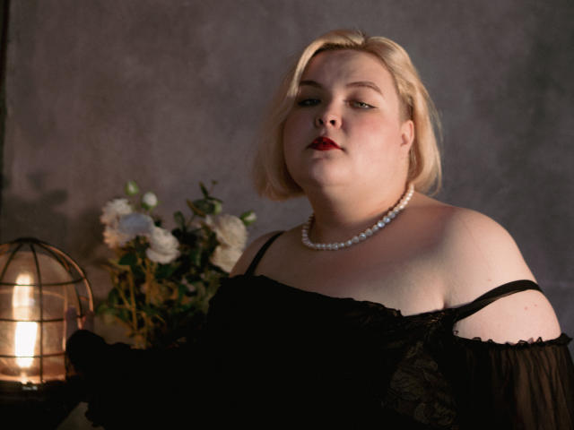 EllaBigBabe with pale skin, blonde bob, red lipstick, black lace off-shoulder top, pearl necklace, looking serious, light from lantern on left, bouquet of white flowers behind.