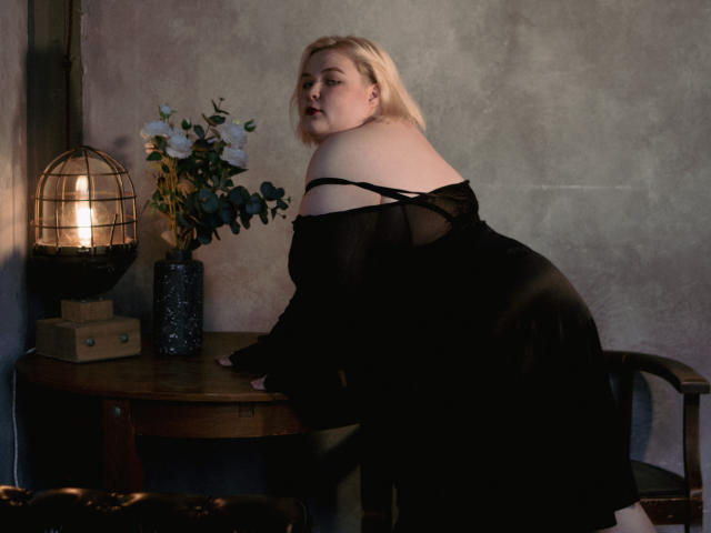 EllaBigBabe with pale skin and blonde hair, wearing a black off-shoulder dress, leaning on a wooden table with a lit lamp and flower vase in a dim room.
