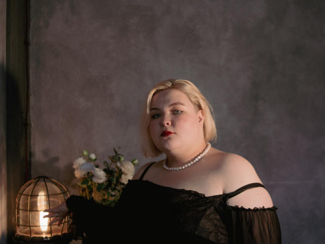 EllaBigBabe with pale skin, blonde bob, red lipstick, black lace off-shoulder dress, pearl necklace, standing, looking serious, lit by cage lamp, bouquet of white flowers.