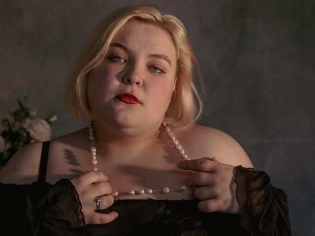 EllaBigBabe has pale skin, blonde hair, and red lipstick. She's holding a pearl necklace, wearing a black sheer top, with tattoos visible on her left shoulder.