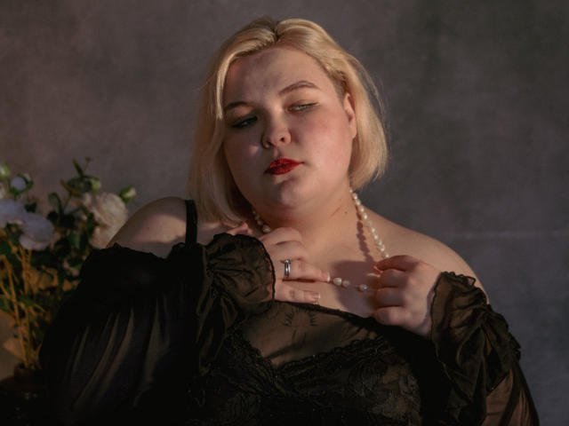 EllaBigBabe has pale skin, blonde bob, red lipstick, black lace dress, pearl necklace, and a ring, looking slightly bored with a soft light on her face.