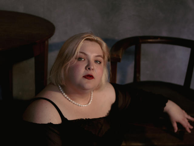 EllaBigBabe, a chubby blonde with pale skin, red lipstick, and a black off-shoulder top, wearing a pearl necklace, leans on a dark wooden chair.