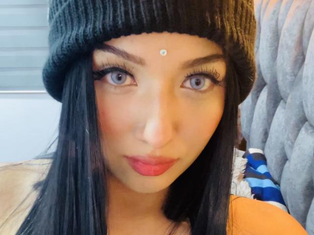 LillyTaylor has black hair, wearing a green beanie, with a diamond-shaped forehead gem, long eyelashes, and red lipstick. Her gray eyes are centered in the selfie.