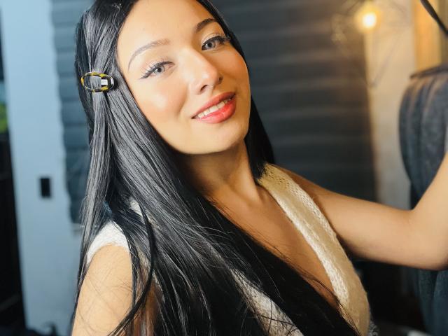 LillyTaylor with long black hair, wearing a white lace top, smiling at the camera, has red lipstick and a gold hair clip, looking cute and sexy.