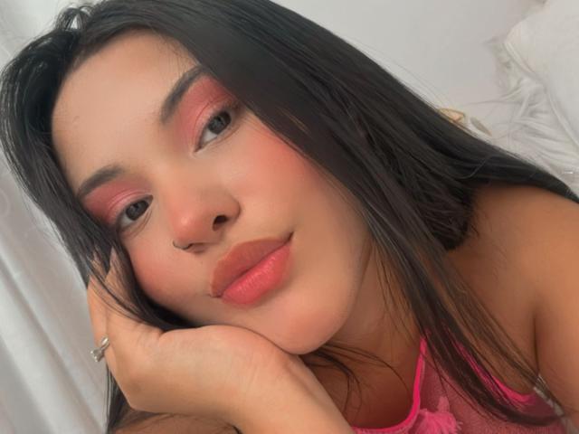 ArianaSmith69 with pink eyeshadow, red lipstick, and nose ring, smiling softly, resting her chin on her hand, wearing a pink lace top, dark hair, and a silver ring.