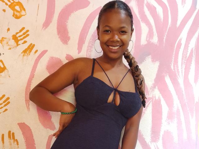 Serayah with dark skin, long braided hair, smiling, wearing a navy blue dress with a keyhole cutout, hoop earrings, green bracelet, hand on hip, showing her tits.