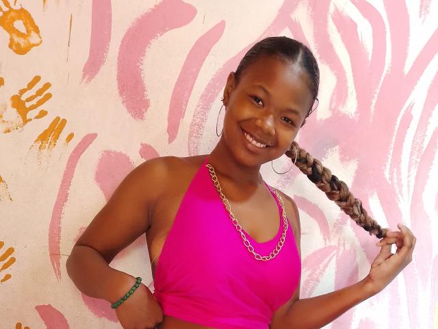 Serayah has dark skin, a braided ponytail, and wears a pink halter top with a gold chain. She's smiling, showing her teeth, with a green bracelet on her wrist.
