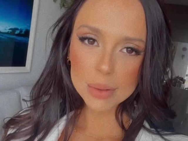 NandaMonteiro has tan skin, long black hair, brown eyes, thick eyelashes, and pink lips. She's wearing a white shirt and a gold necklace. Her ass is not visible.